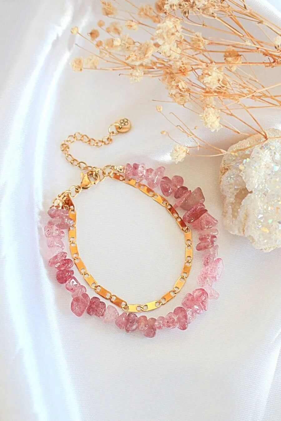 Bracelet with pink gemstones and gold chain on a white surface with dried flowers.