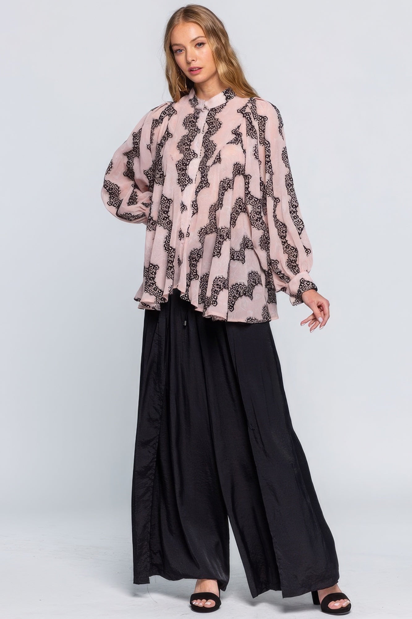 Woman wearing a pink blouse with black pattern and black wide-leg pants on a white background