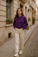 Woman in a purple shirt and beige pants standing on a street with buildings and plants in the background.