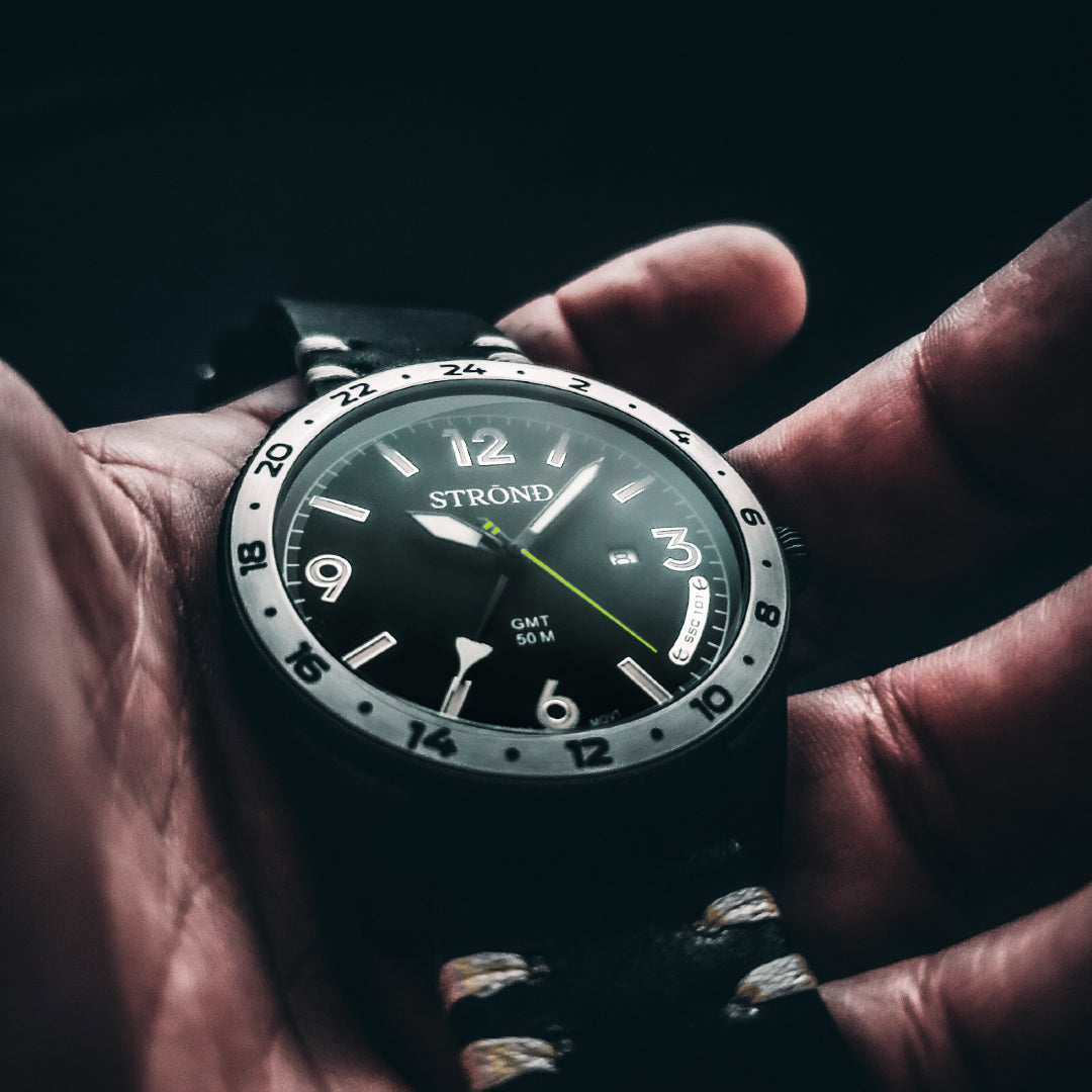 Hand holding a Strond watch with a dark face and leather strap against a black background