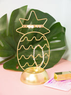 Gold pineapple-shaped jewelry holder with a bracelet on a pink surface with a green leaf background
