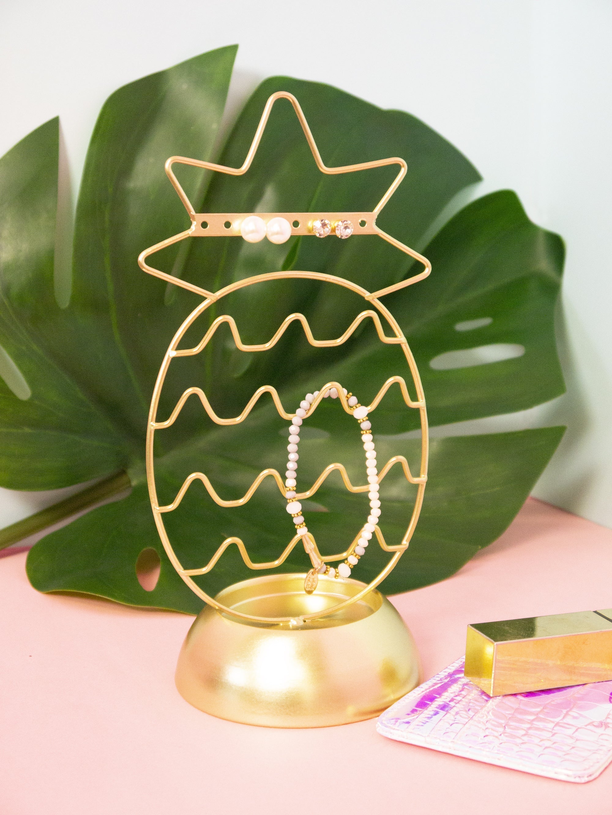 Gold pineapple-shaped jewelry holder with a bracelet on a pink surface with a green leaf background