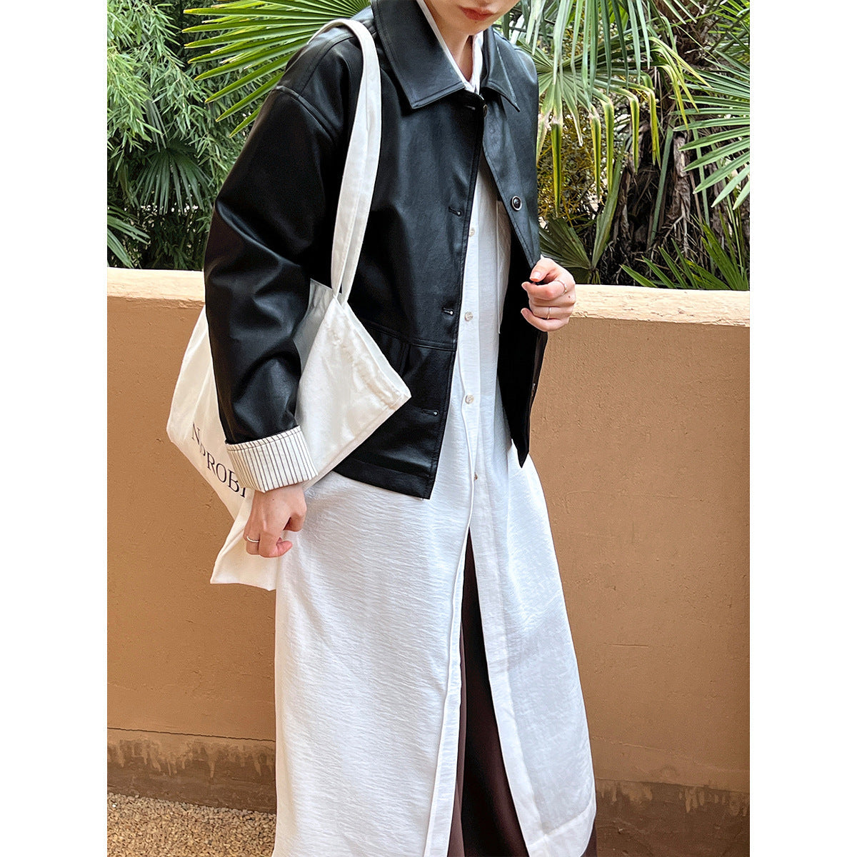 Person wearing a black leather jacket over a white shirt and pants, holding a beige bag with text.