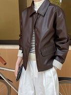 Person wearing a brown leather jacket with a striped shirt and white pants, standing next to a bicycle.