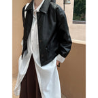 Person wearing a black jacket over a white shirt and brown pants against a beige wall.