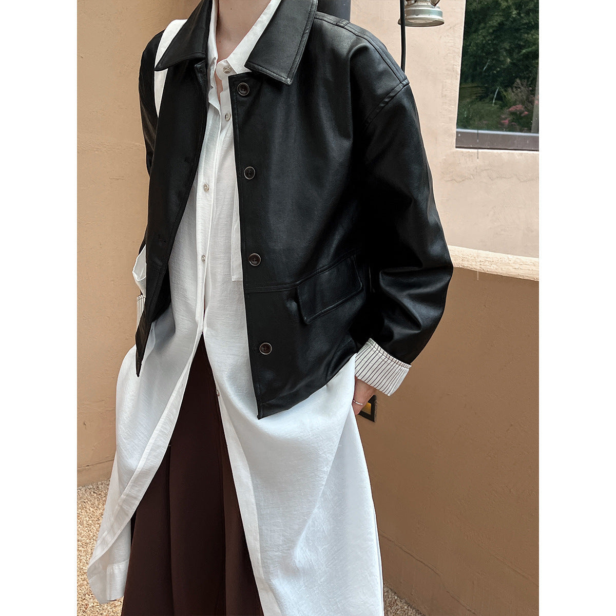 Person wearing a black jacket over a white shirt and brown pants against a beige wall.