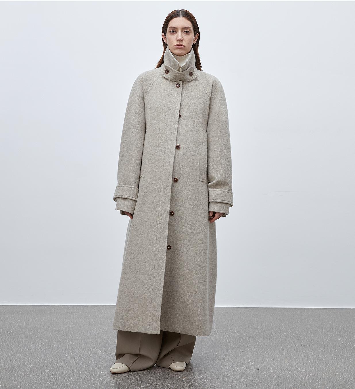 Person wearing a long beige coat against a plain background