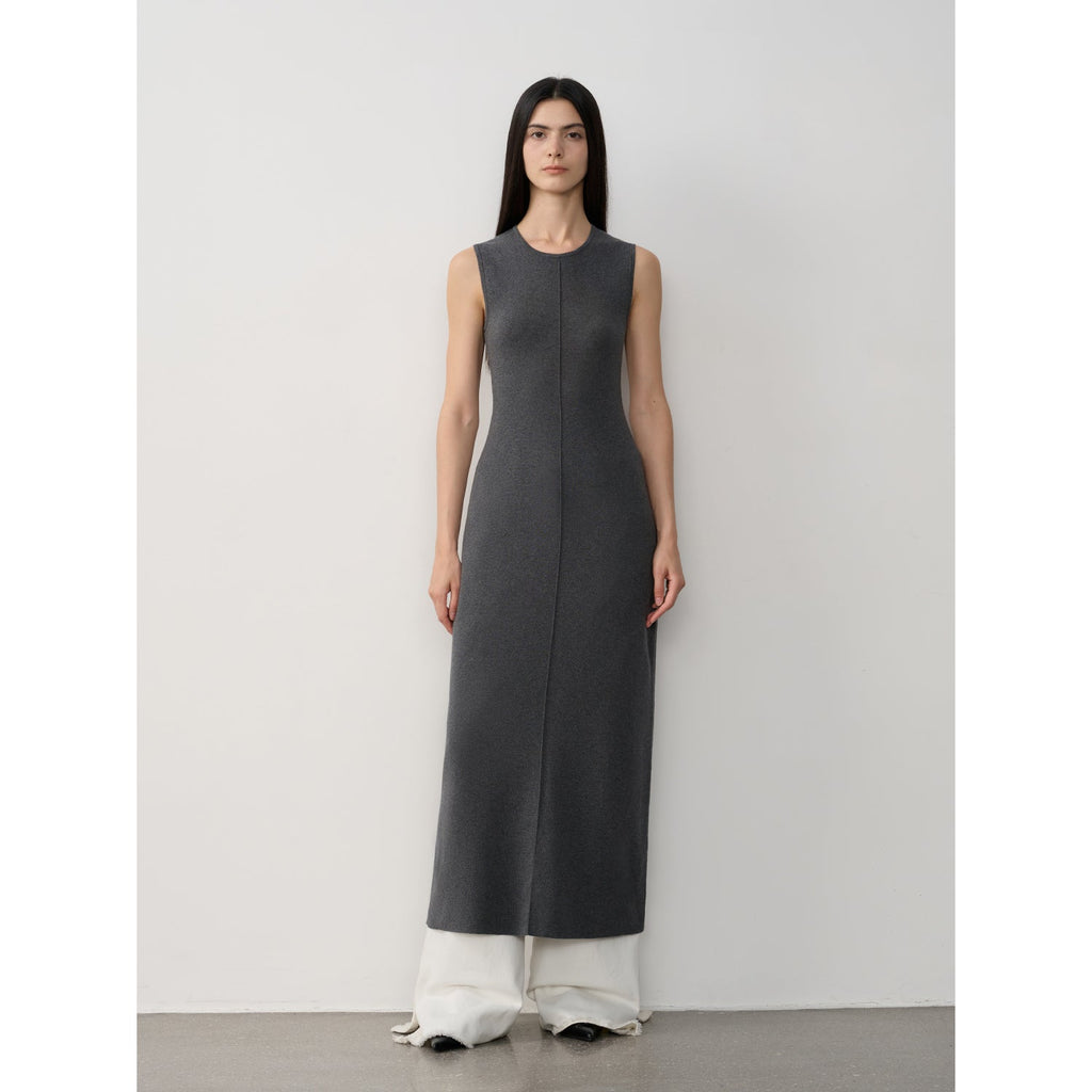 Woman wearing a long dark gray dress with white pants against a plain background