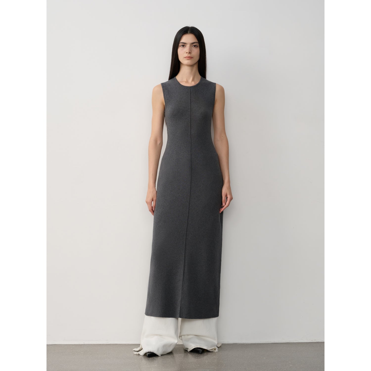 Woman wearing a long dark gray dress with white pants against a plain background