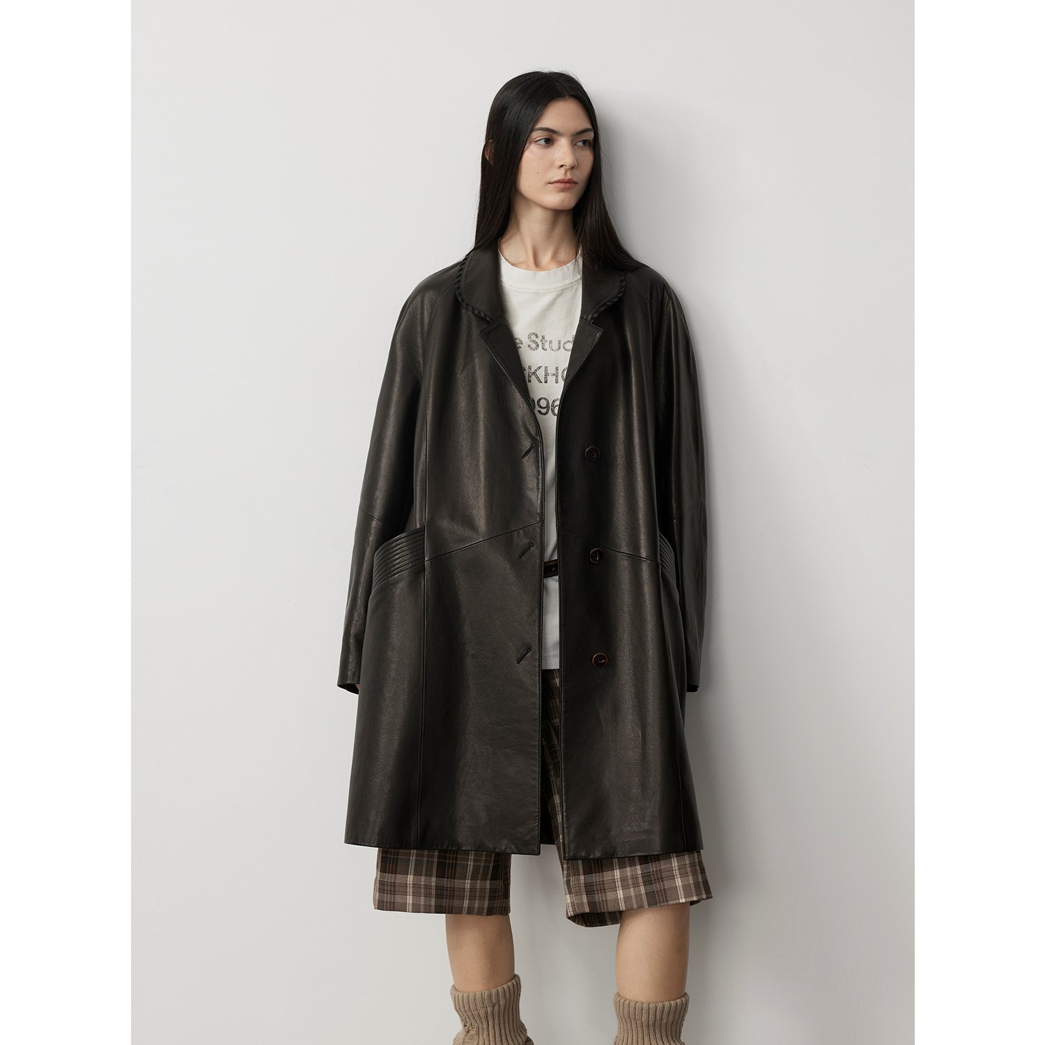 Person wearing a long dark coat over a white shirt with text, plaid skirt, and brown shoes on a light gray background.