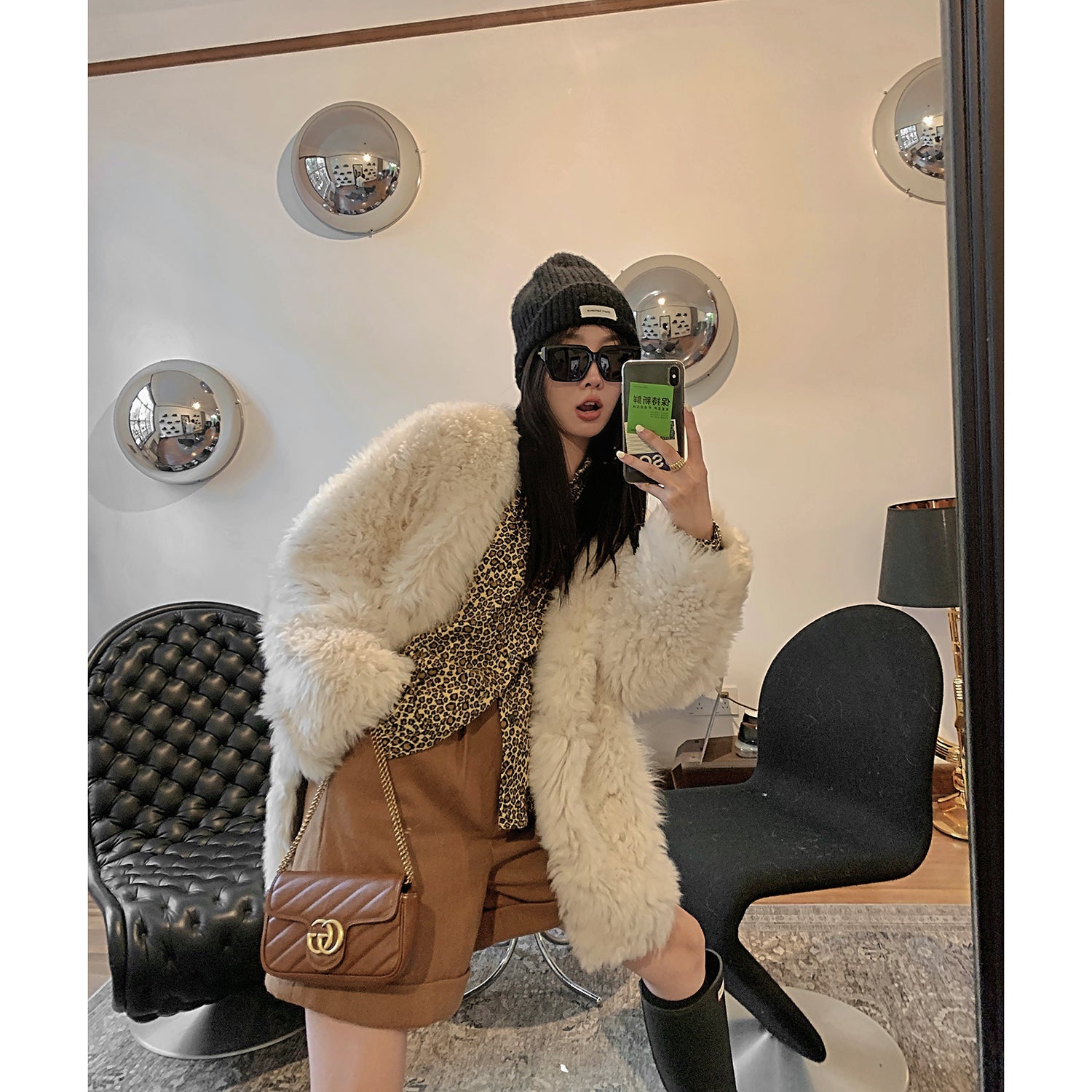 Person taking a mirror selfie wearing a white fur coat, brown skirt, and black boots in a stylish room.