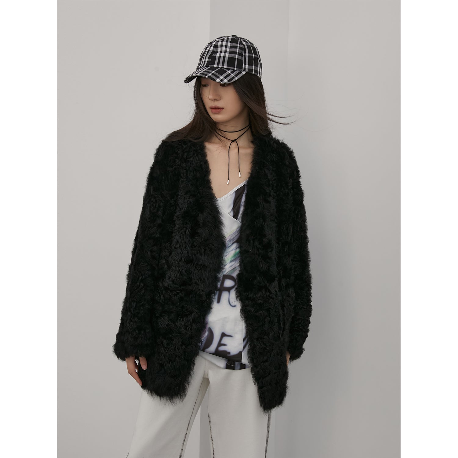 Person wearing a black fluffy cardigan, white pants, and a plaid cap against a plain background