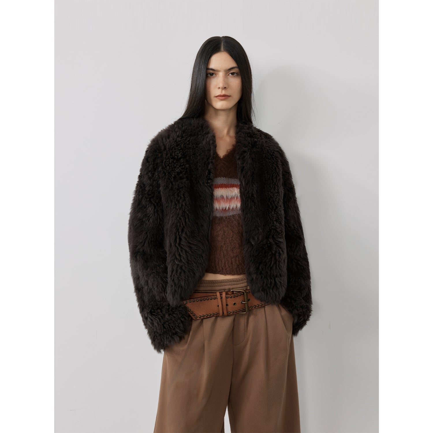 Person wearing a dark brown fur coat over a patterned sweater and brown pants on a light gray background