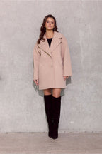 Woman wearing a beige coat and black knee-high boots against a gray concrete wall.