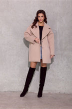 Woman wearing a beige coat and black knee-high boots against a gray concrete wall.