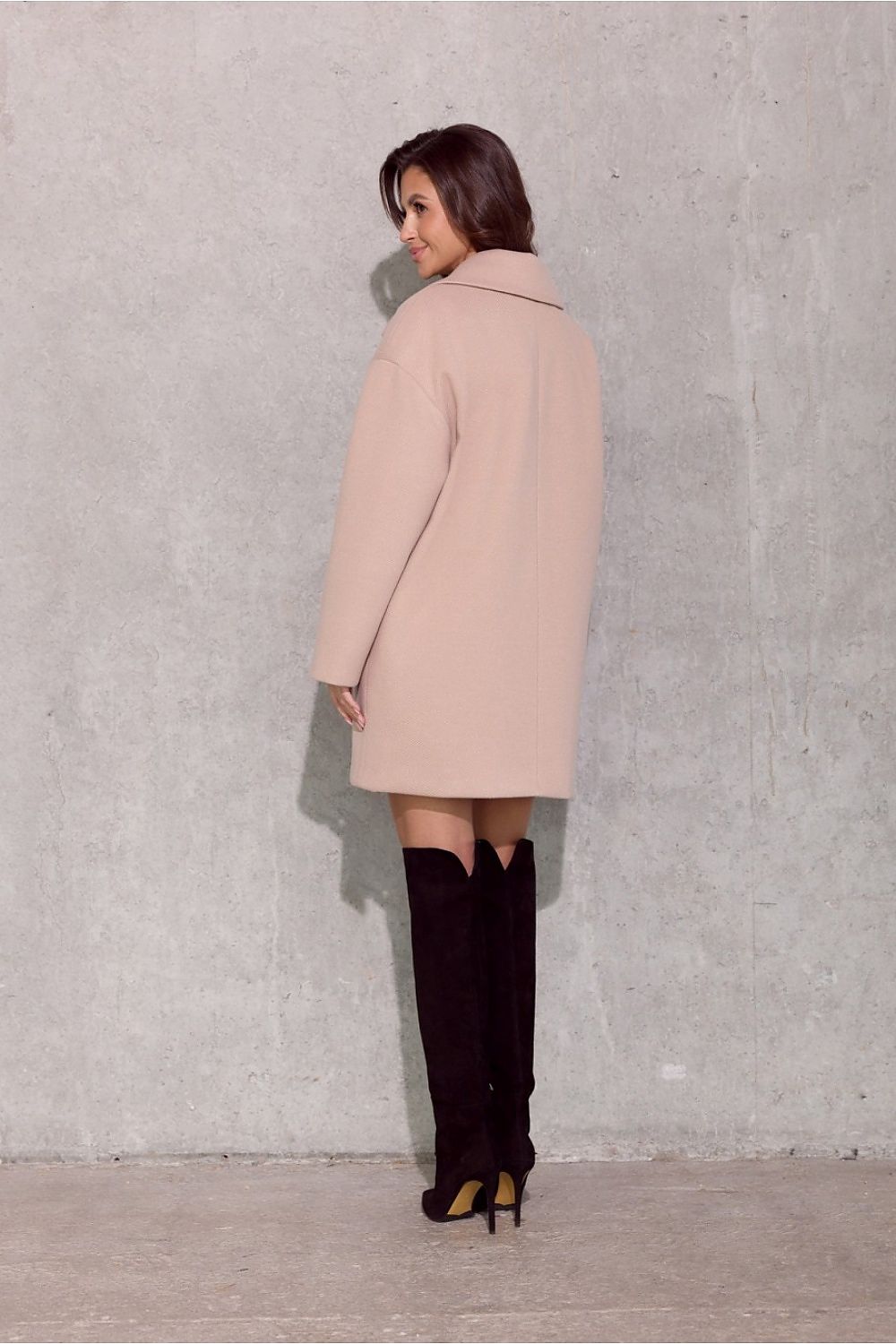 Woman wearing a light pink coat and black knee-high boots against a gray wall.