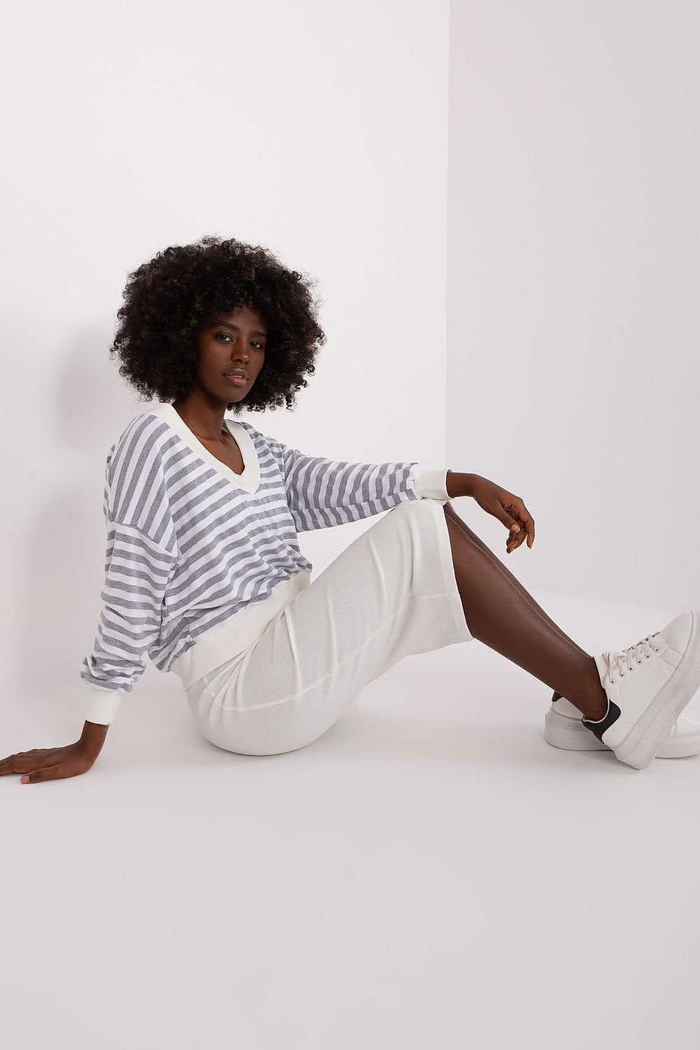 Person wearing a striped shirt and white pants sitting on a white floor.
