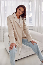 Woman wearing a beige coat and light blue jeans sitting on a white couch in a bright room.