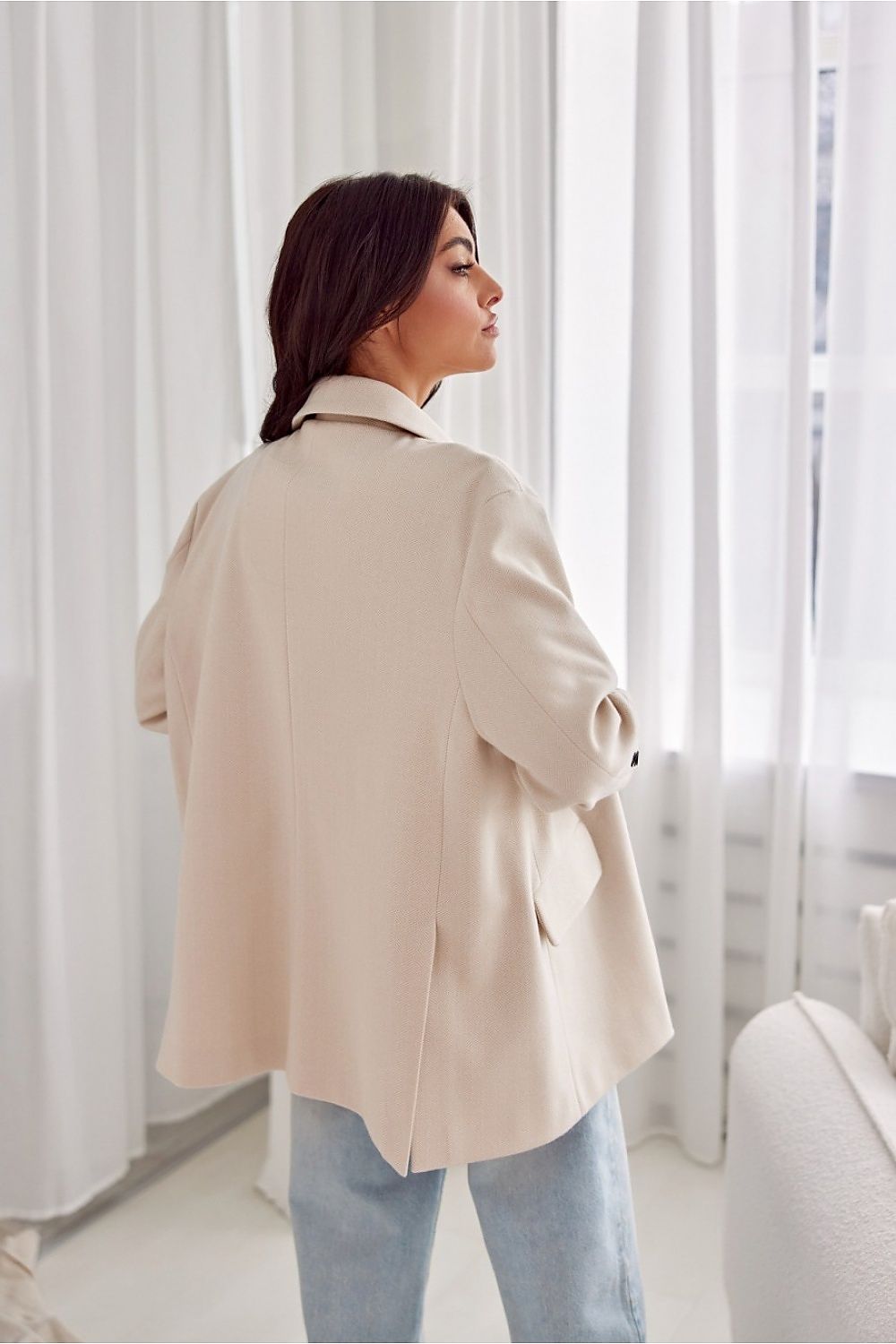 Person wearing a beige coat standing in a bright room with white curtains.