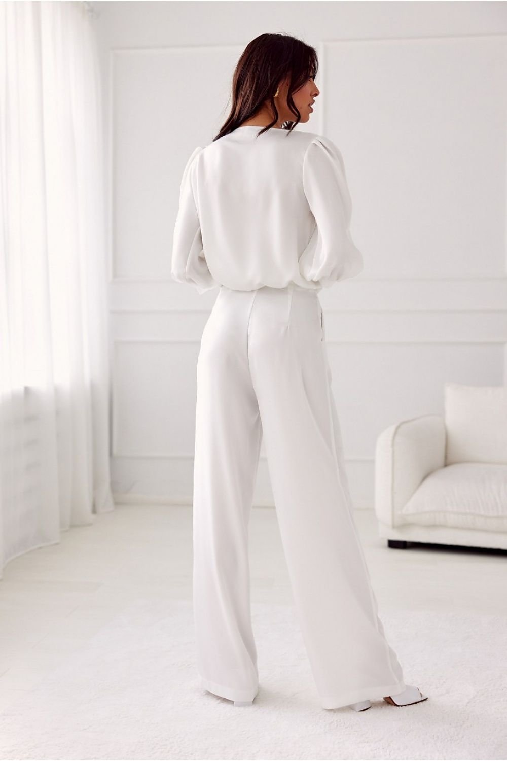Woman in a white outfit standing in a minimalistic room with white walls and furniture.