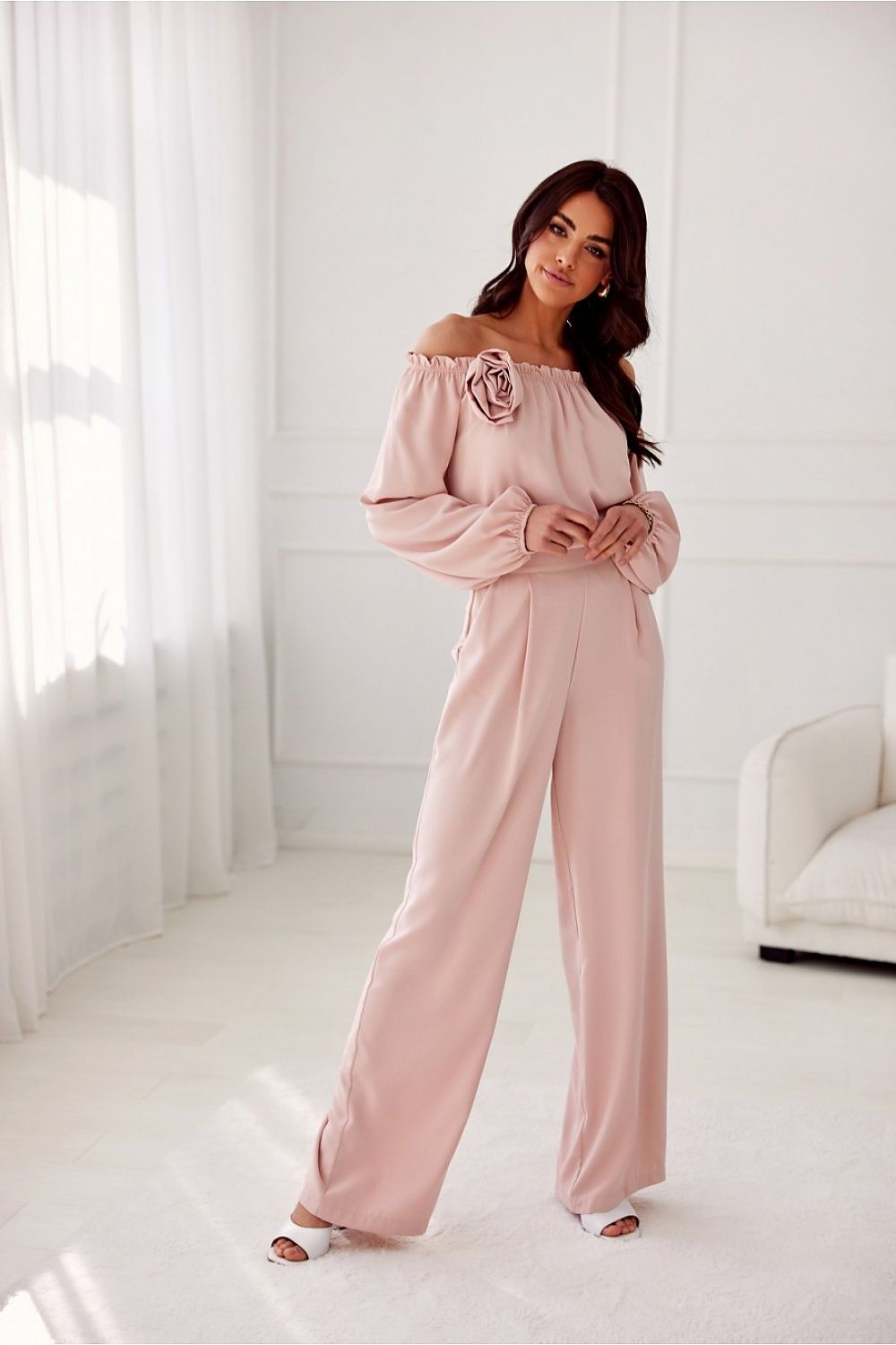 Woman wearing a pink off-shoulder top and high-waisted pants in a bright room.