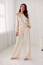 Woman wearing a beige off-shoulder top and wide-leg pants in a bright room.