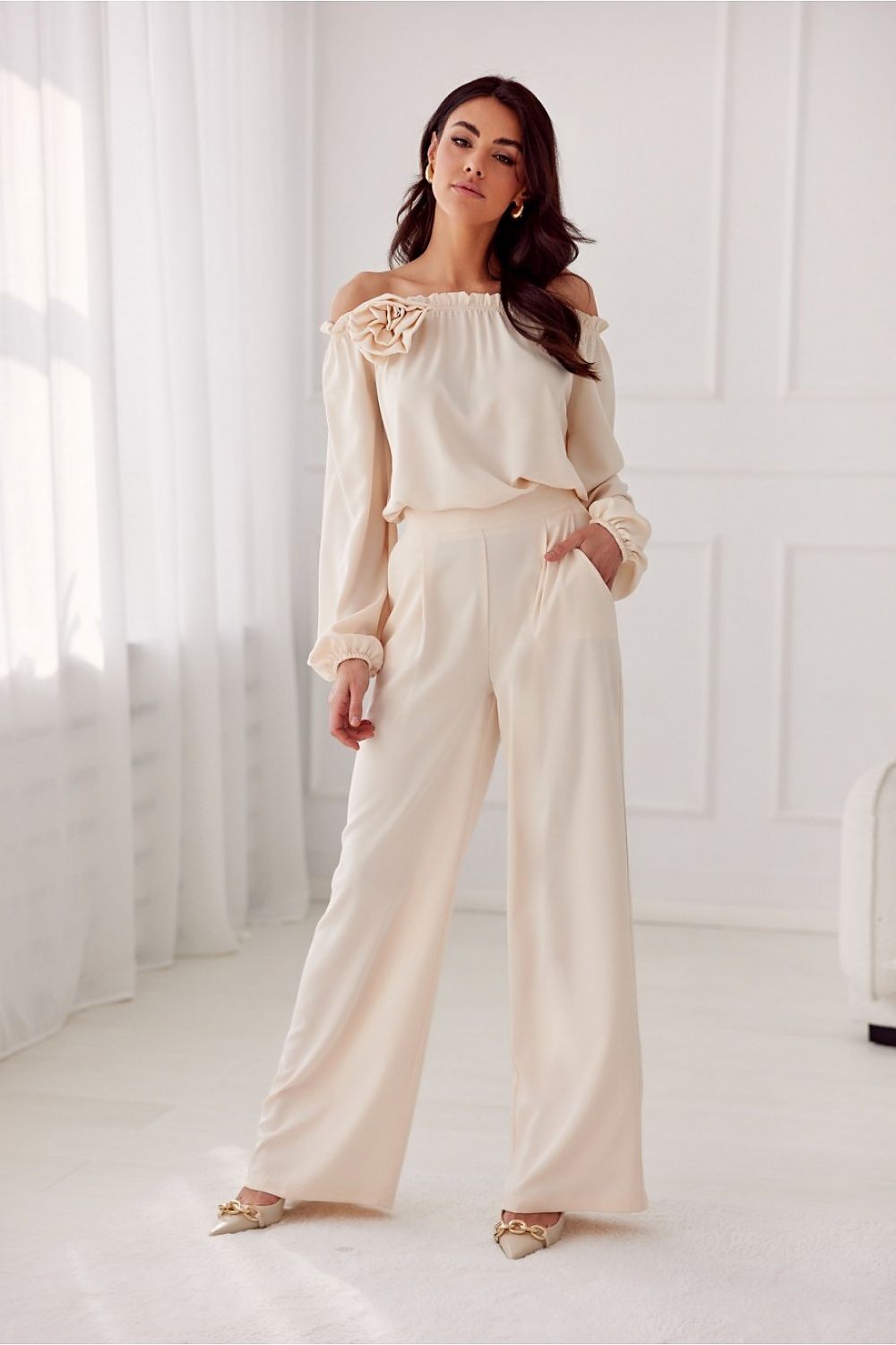 Woman wearing a beige off-shoulder top and wide-leg pants in a bright room.