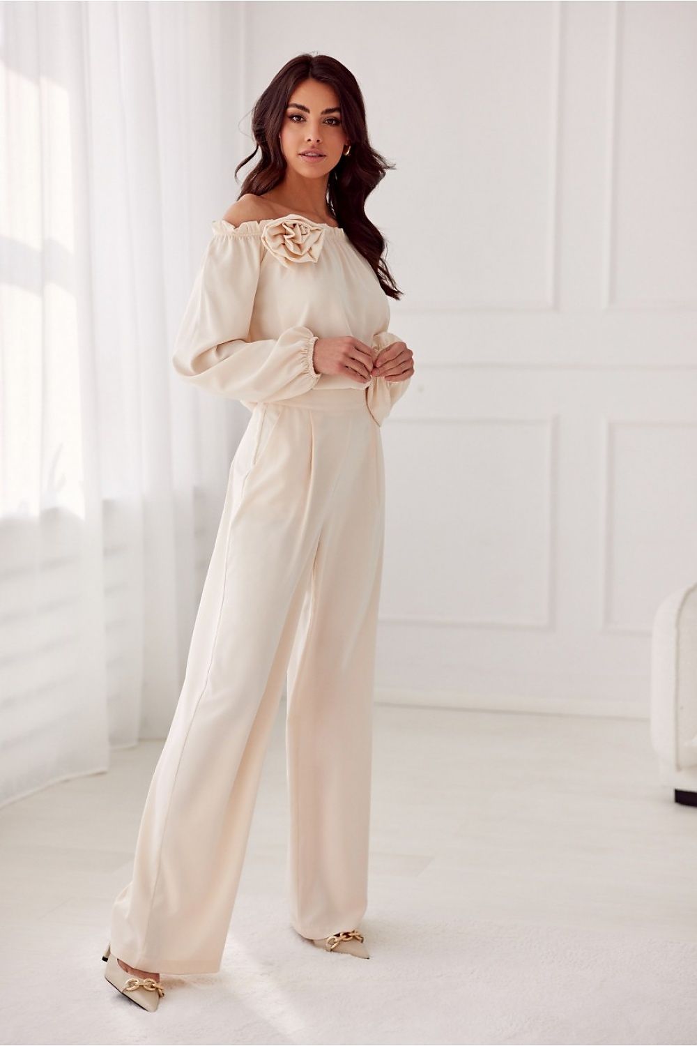 Woman wearing a cream-colored off-shoulder top and wide-leg pants in a bright room.