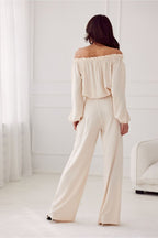 Woman wearing a cream off-shoulder top and wide-leg pants in a minimalistic room.
