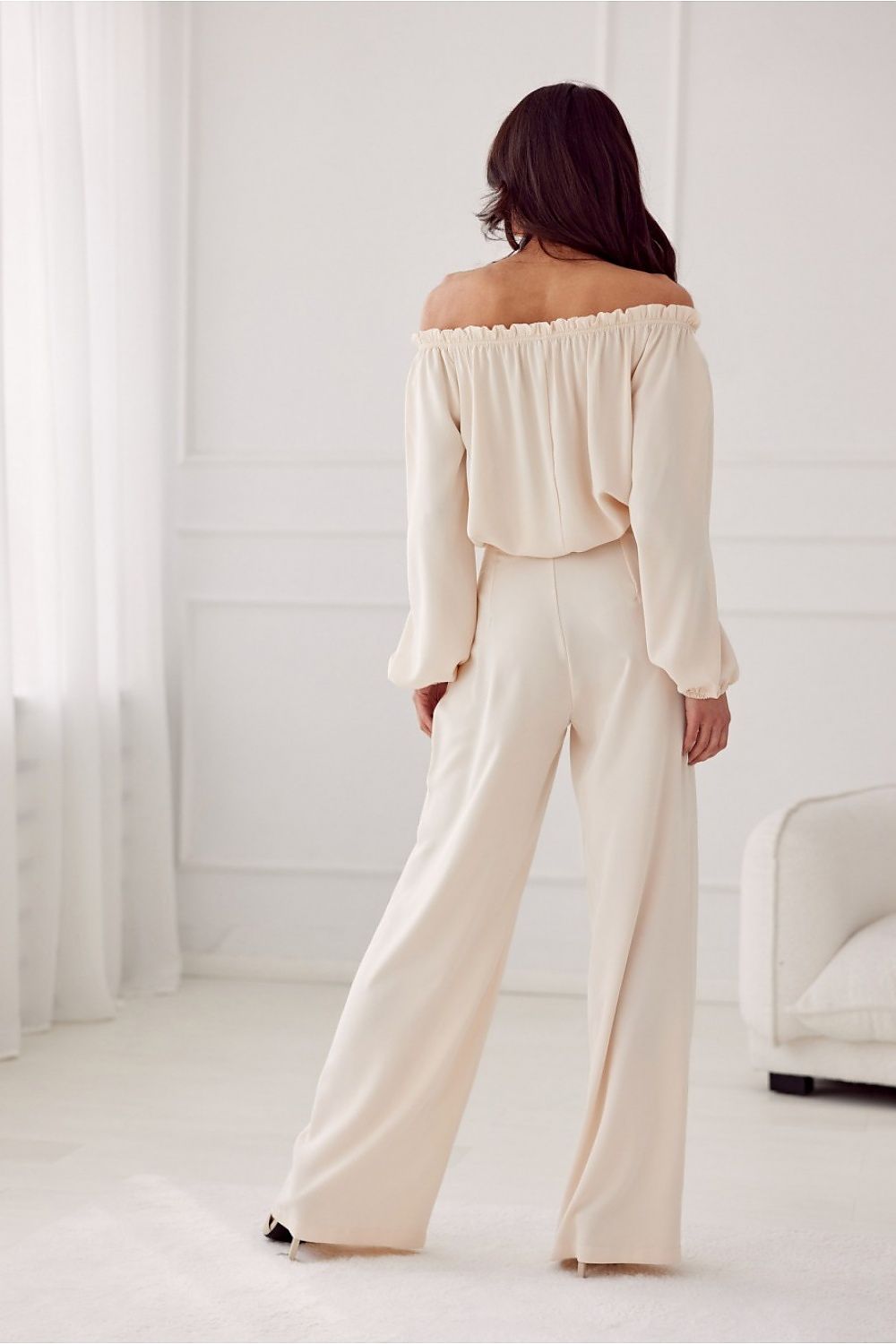 Woman wearing a cream off-shoulder top and wide-leg pants in a minimalistic room.
