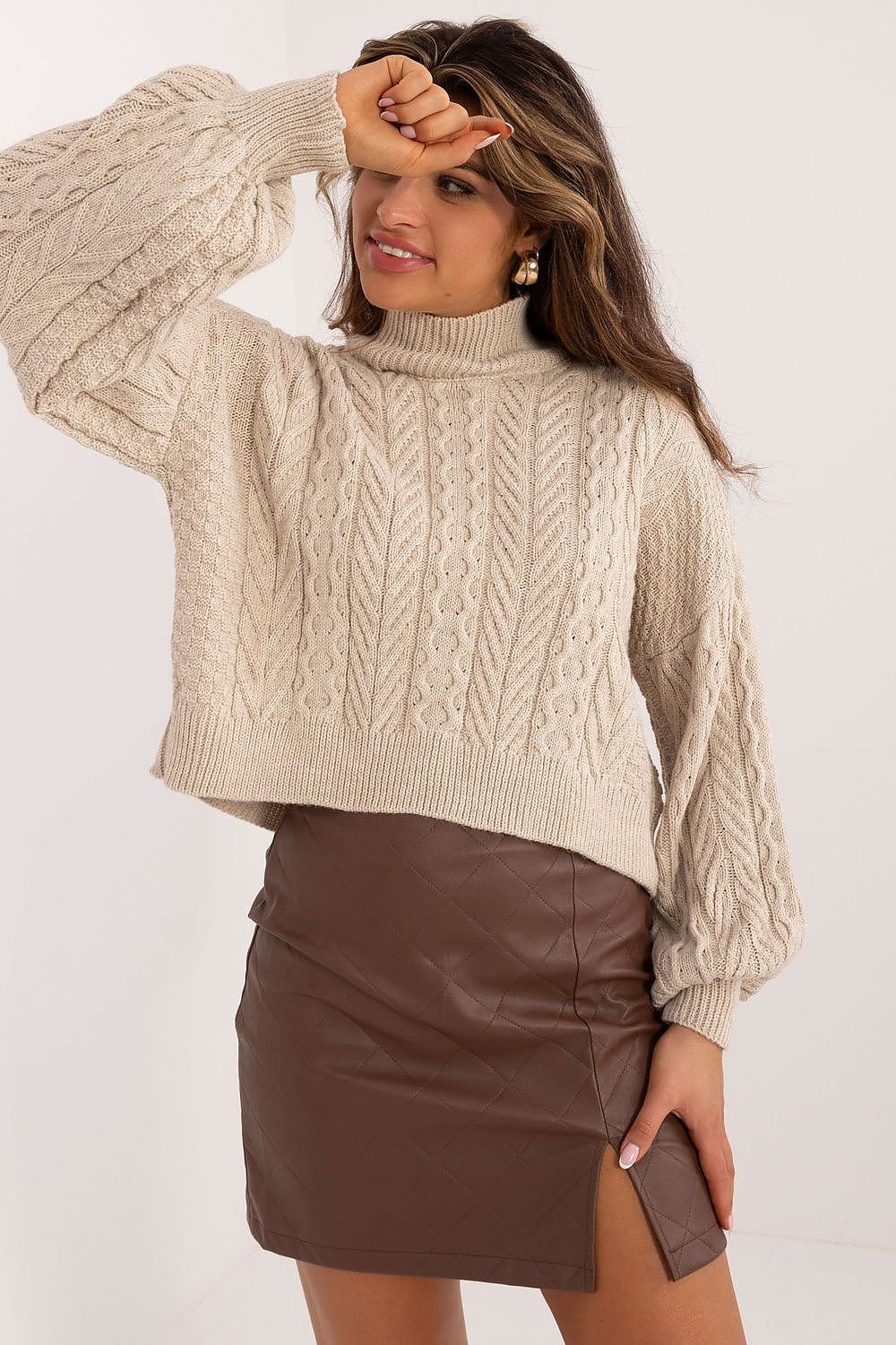 Woman wearing a beige cable knit sweater and brown leather skirt on a white background
