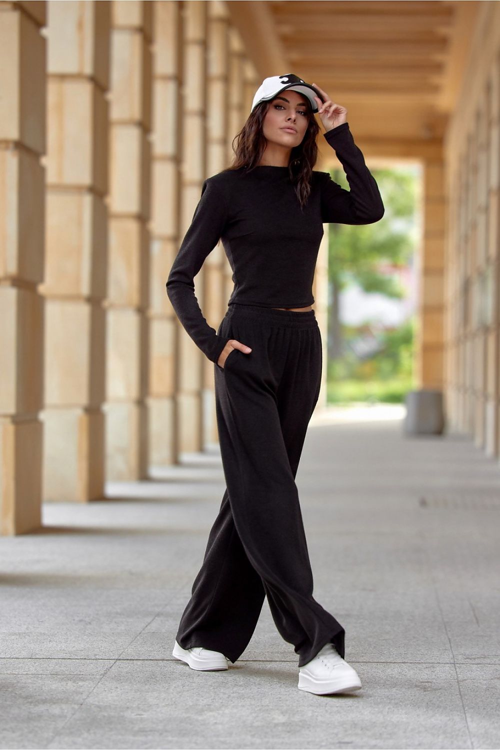Woman in black outfit with white sneakers standing in a sunlit outdoor setting.
