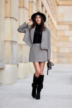 Woman in gray coat and skirt with black boots and hat standing in front of a building.