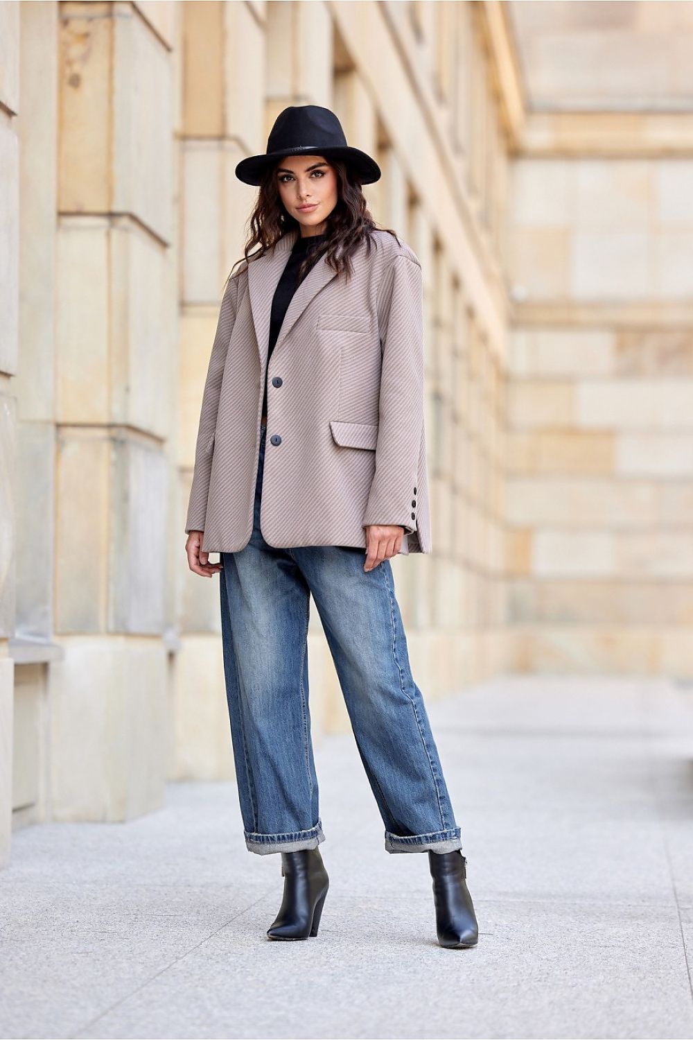 Woman wearing a beige blazer, black hat, blue jeans, and black boots standing in front of a stone wall.