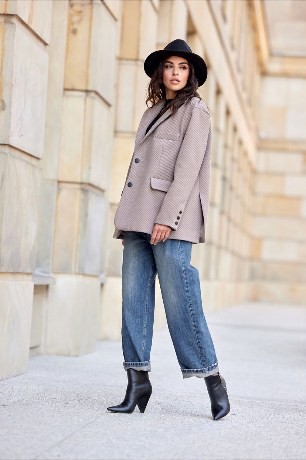 Woman wearing a beige coat, black hat, blue jeans, and black boots standing in front of stone architecture.