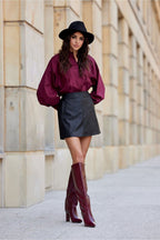 Woman wearing a burgundy blouse, black skirt, and high boots against a stone wall.