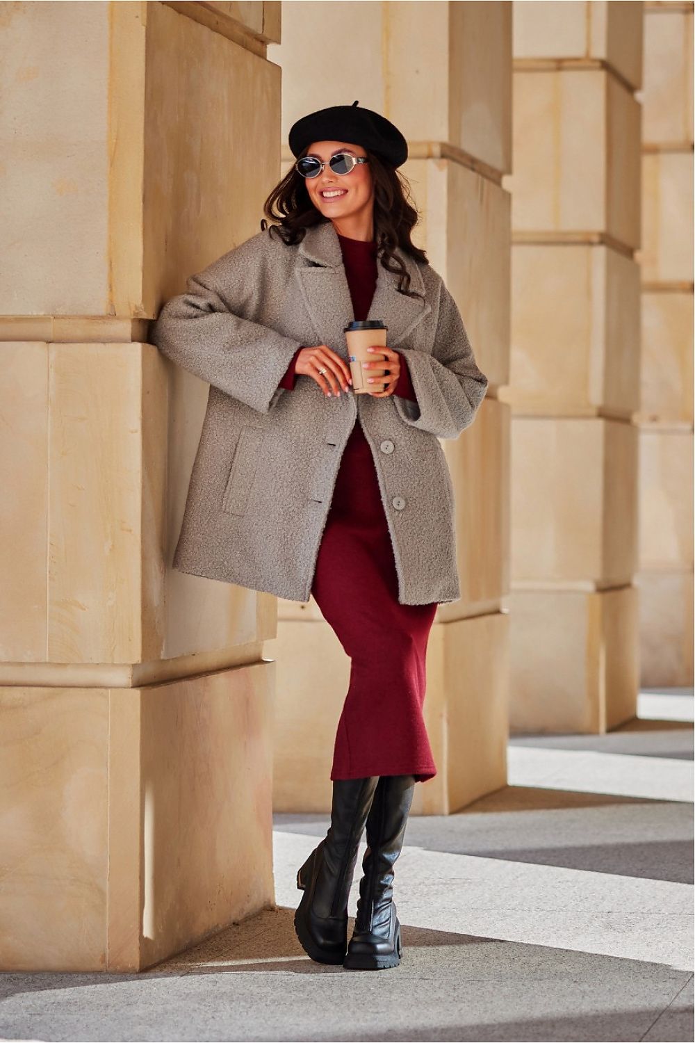 Woman in a gray coat and red dress standing against a stone wall.