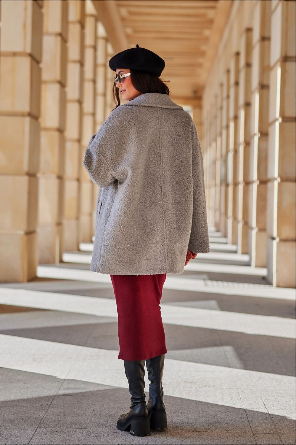 Person wearing a gray coat and red pants walking through a sunlit architectural hallway.