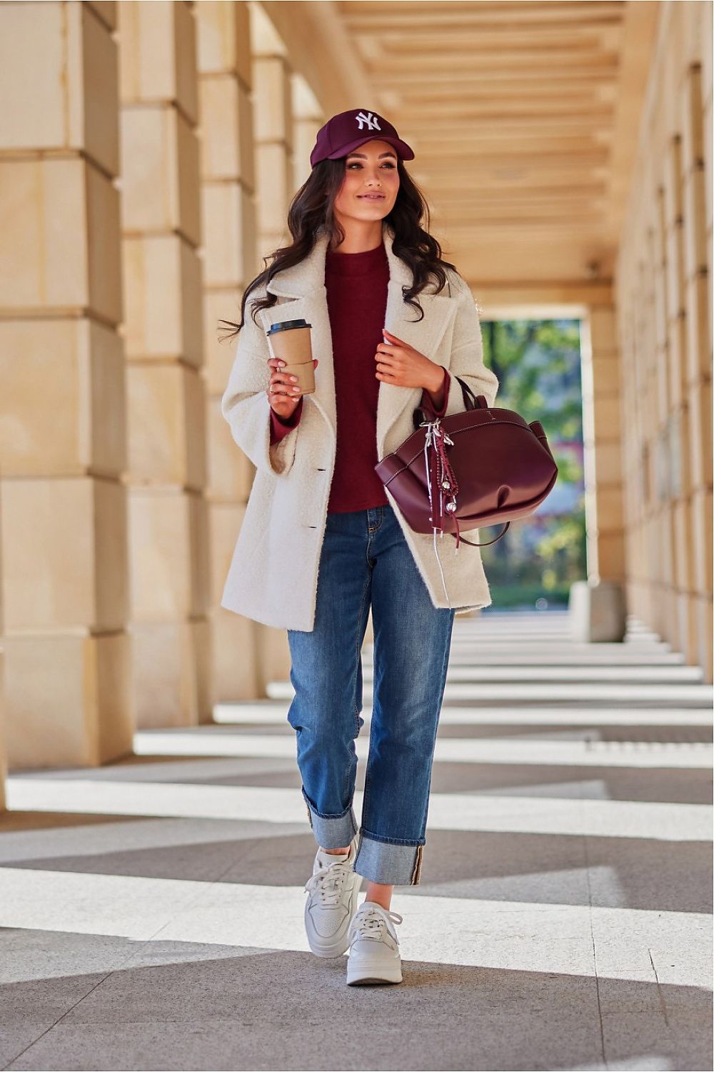 Woman walking outdoors holding a coffee cup and a handbag, wearing a white coat, red sweater, blue jeans, and a purple cap.