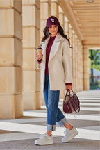 Woman walking outdoors wearing a beige coat, blue jeans, and a purple cap, holding a coffee cup and handbag.