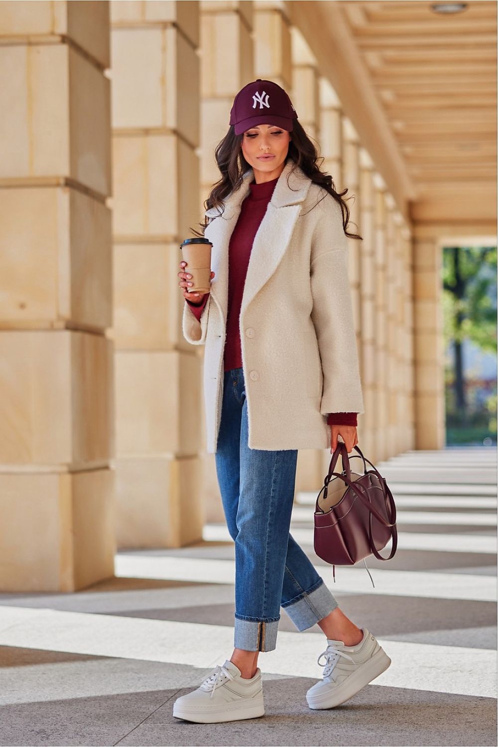 Woman walking outdoors wearing a beige coat, blue jeans, and a purple cap, holding a coffee cup and handbag.