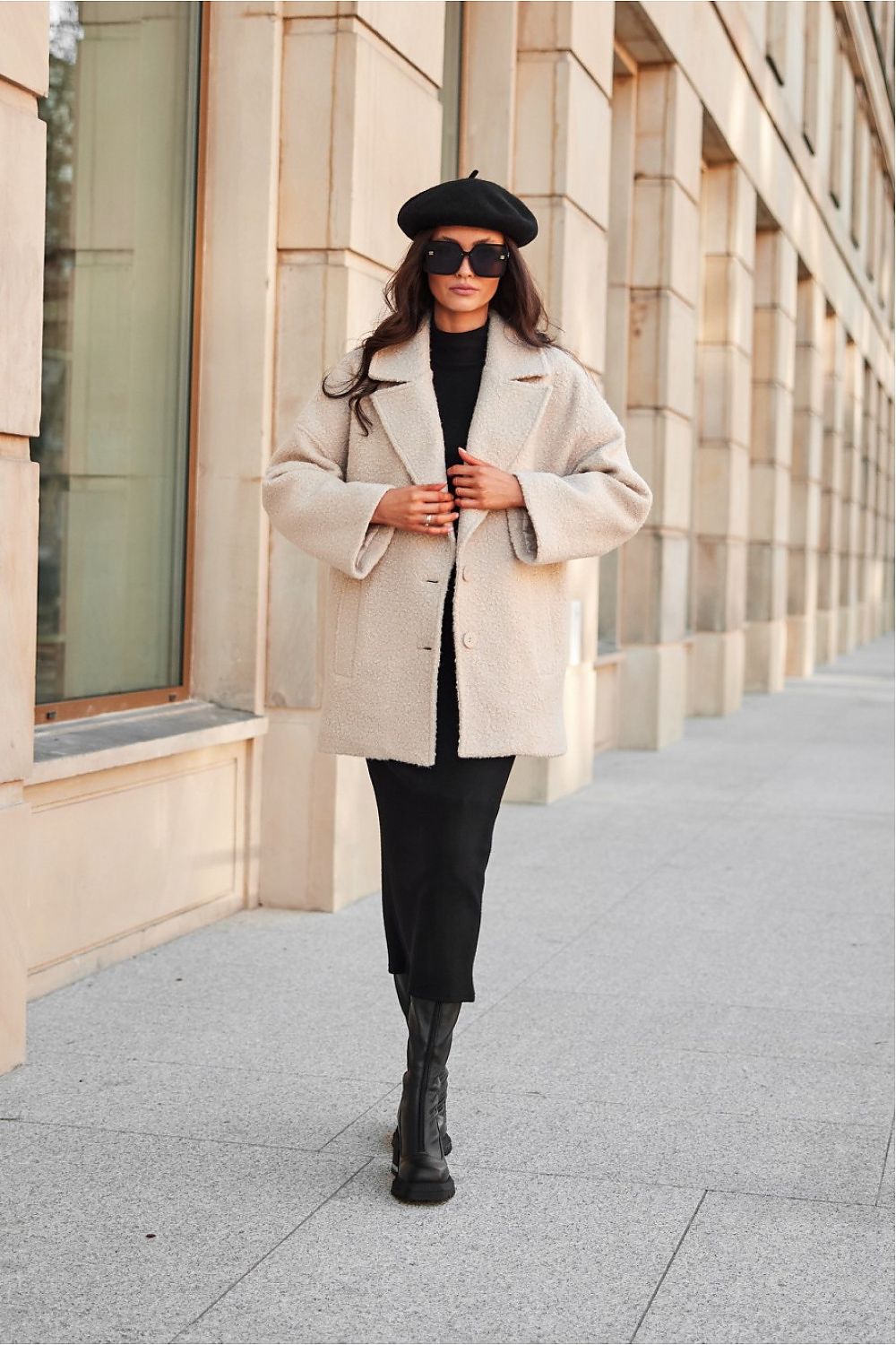 Woman in a beige coat, black beret, and sunglasses walking on a sidewalk.