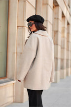 Woman wearing a beige coat, black beret, and sunglasses standing against a building.