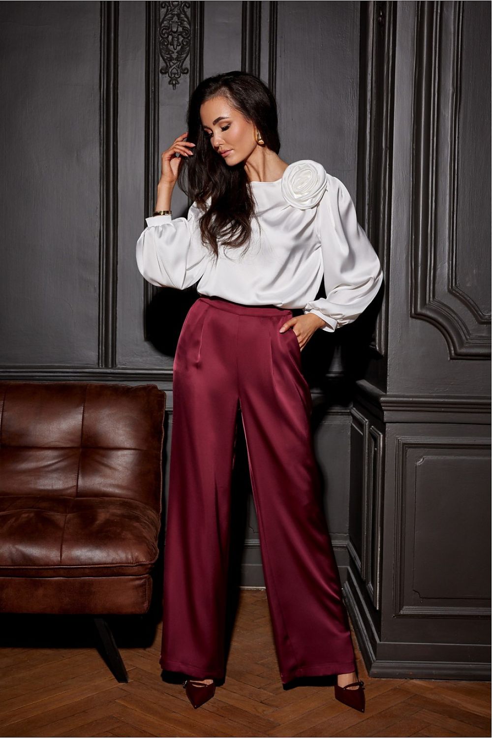 Woman in a white blouse and burgundy pants standing in a room with dark paneling and a brown leather couch.