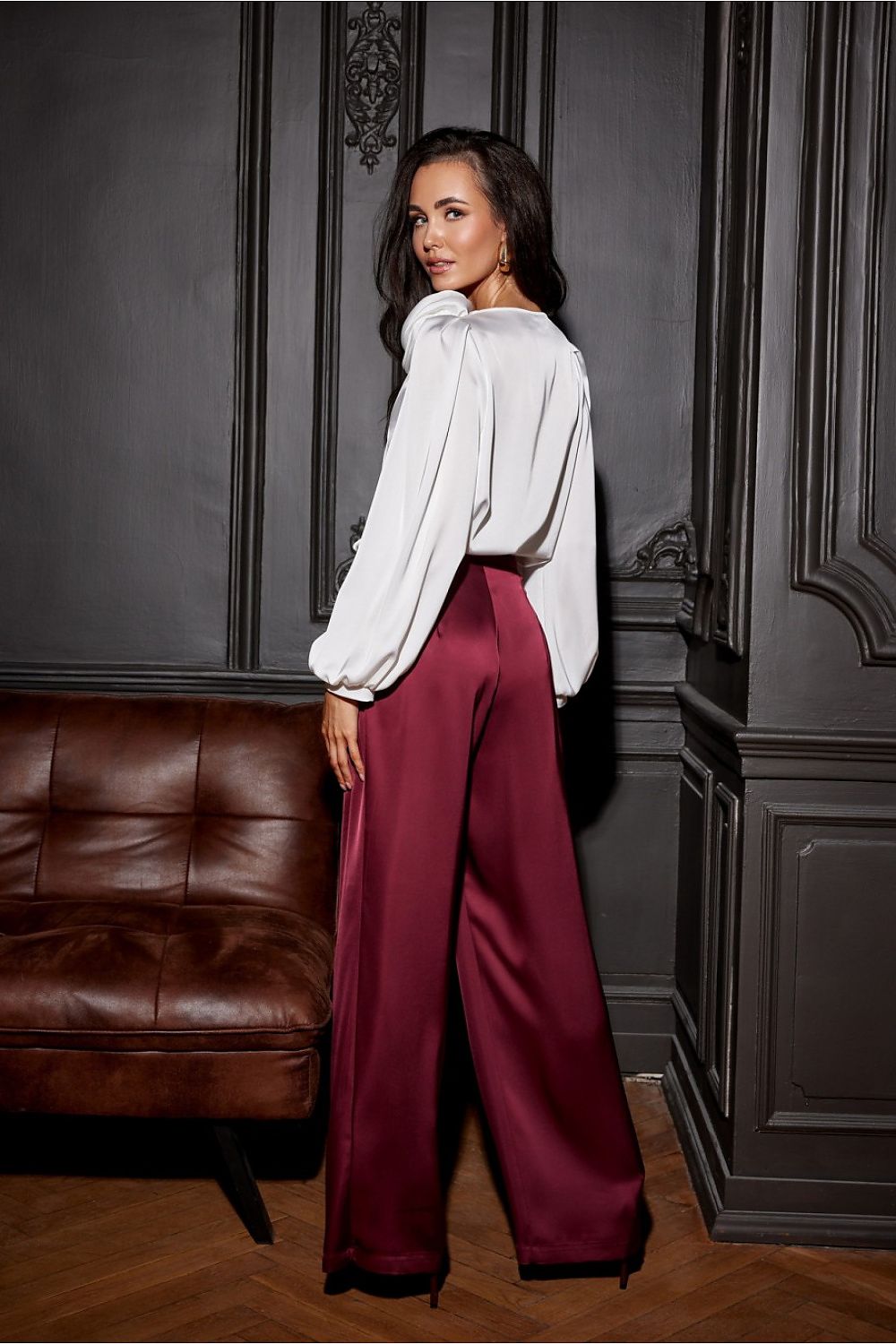 Woman in a white blouse and red pants standing in a room with dark paneling and a brown leather couch.