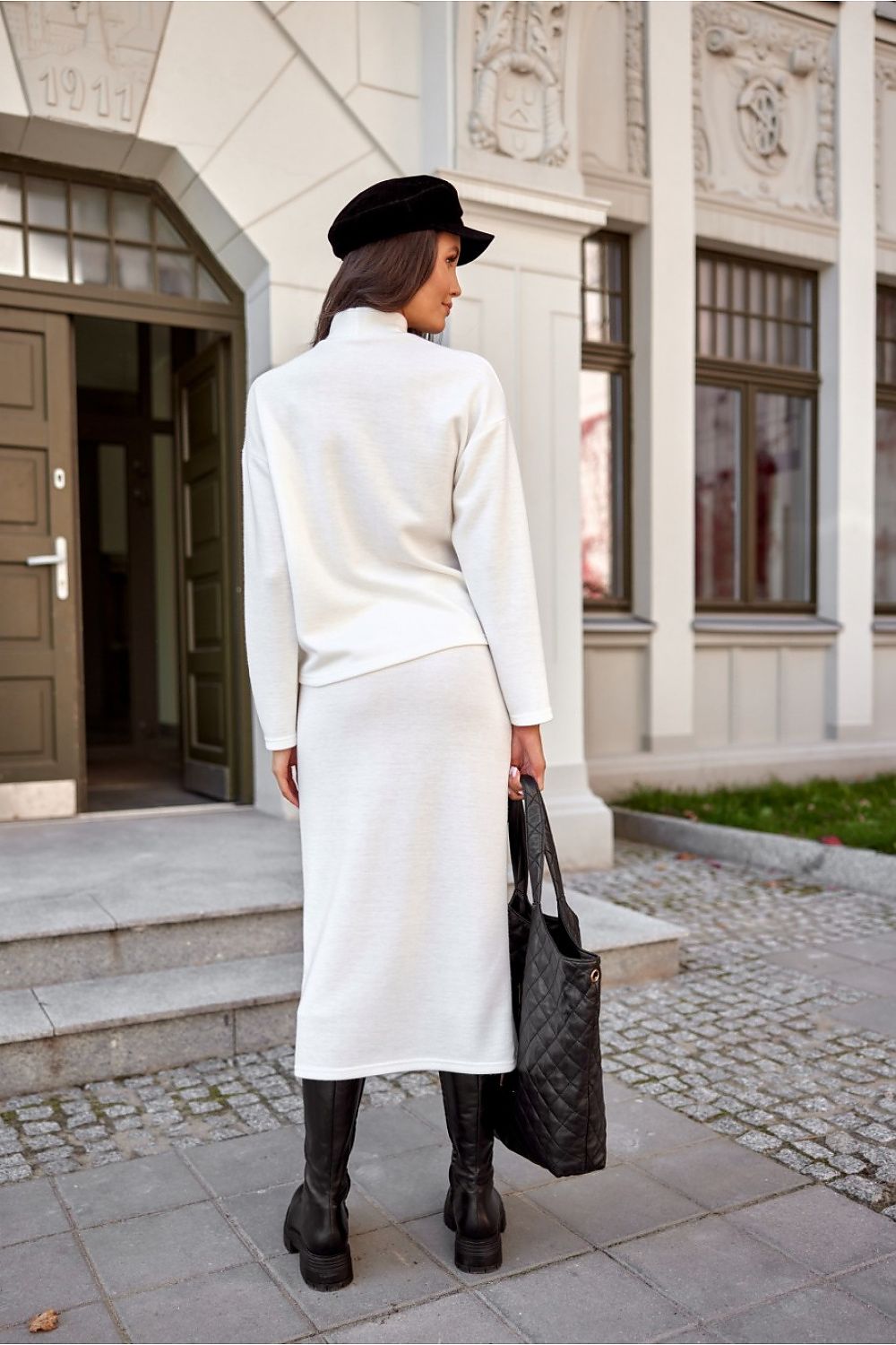 Woman in a white outfit with a black hat and bag walking outside a building.