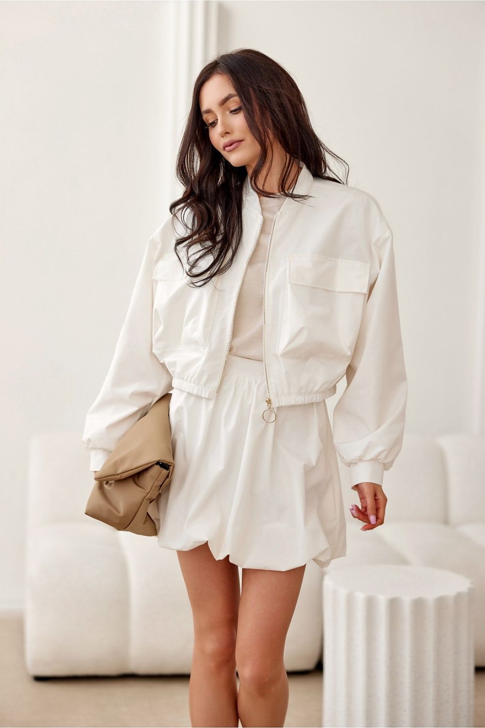 Woman wearing a white outfit with a beige bag in a minimalistic room.