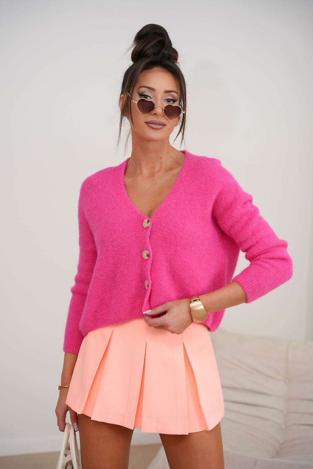 Woman wearing a bright pink cardigan and peach skirt with sunglasses on a plain background