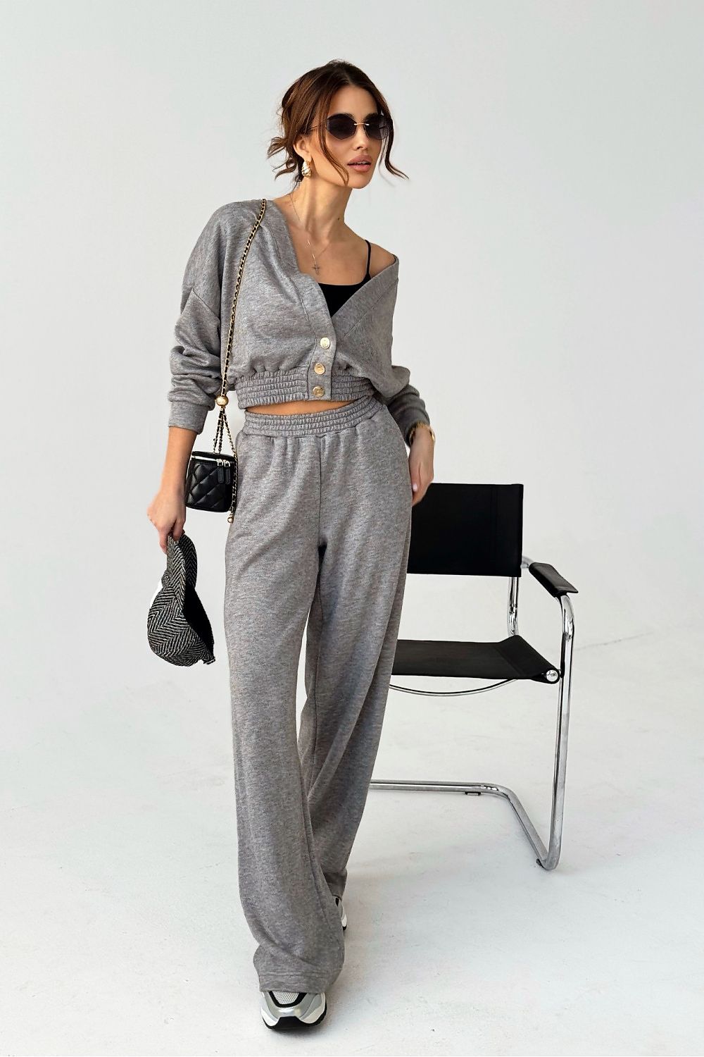Woman in gray outfit standing next to a black chair on a white background