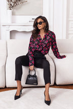 Woman in a stylish outfit with a floral blouse and black pants sitting on a white couch.